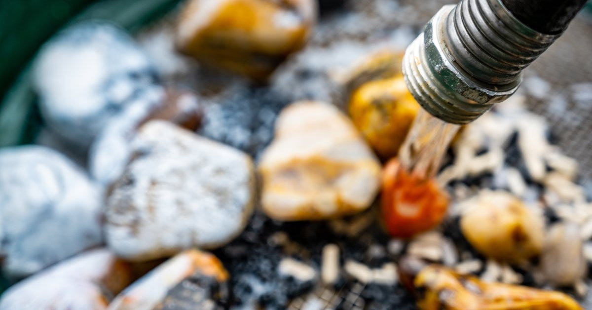 A hose rinses stones on a metal screen in a rock tumbler. Water pours over all the rocks, which are out of focus.