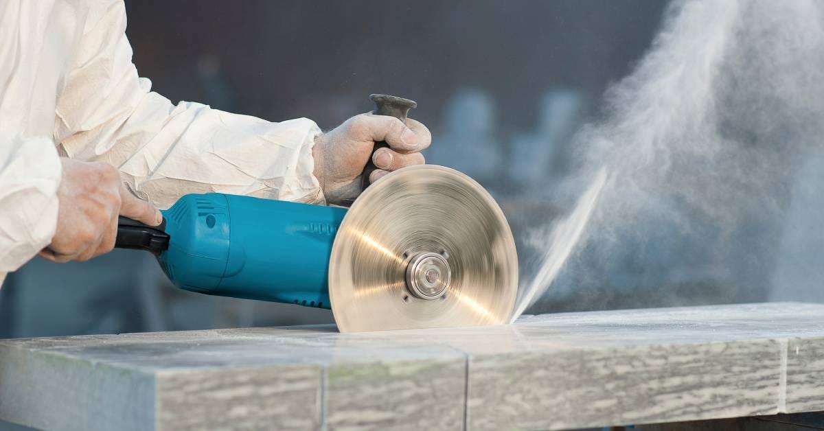 A man wearing a protective white uniform. He holds a blue grinder with a diamond blade while cutting a flat stone.