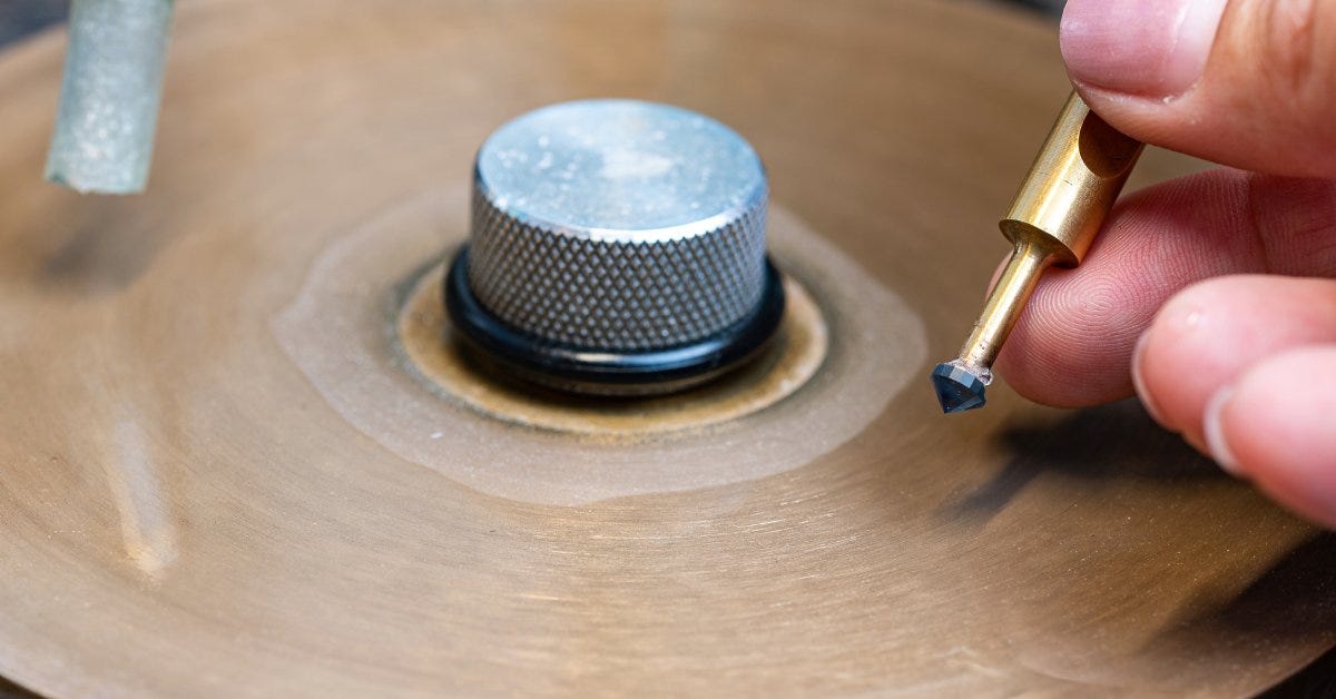 A lapidary tool holds a blue sapphire on its tip as a person prepares to brush it against a metal faceting machine.