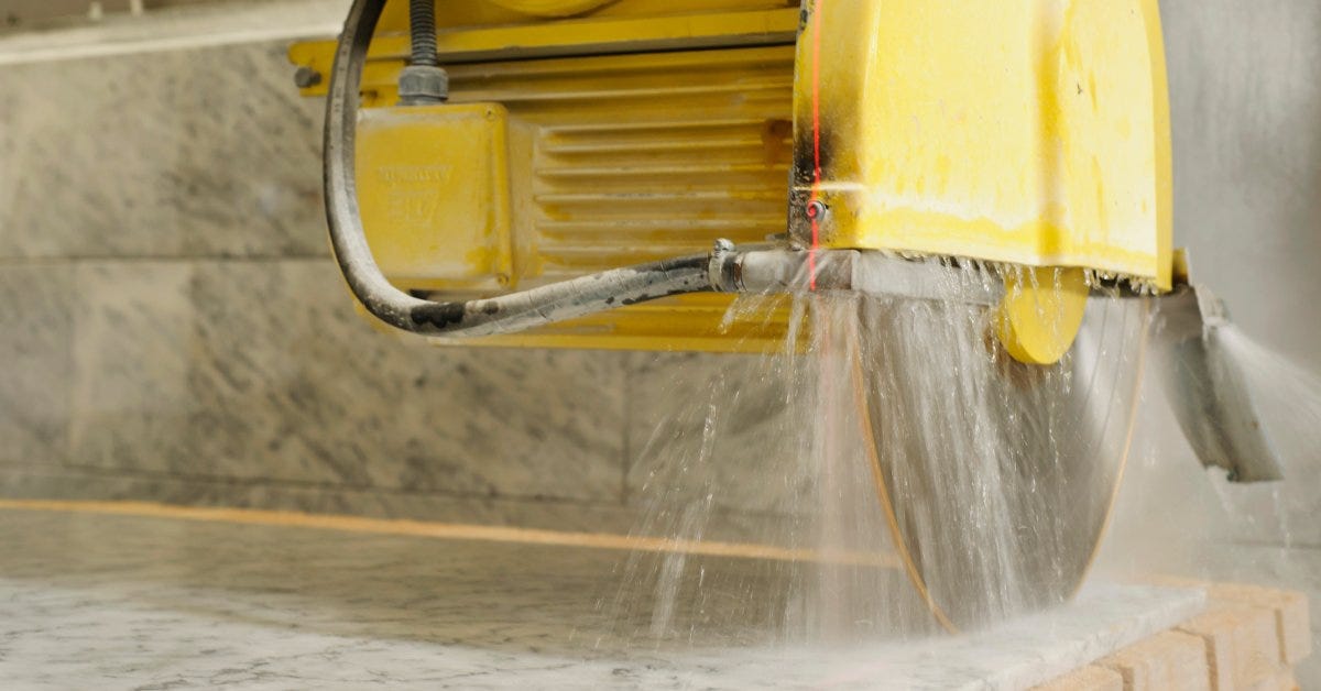 A yellow slab saw cuts through the surface of a marble panel. Water sprouts onto the material as it moves through it.