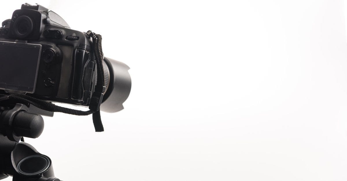 A black, professional DSLR camera sits on a tripod at a slightly downward angle against a bright white background.