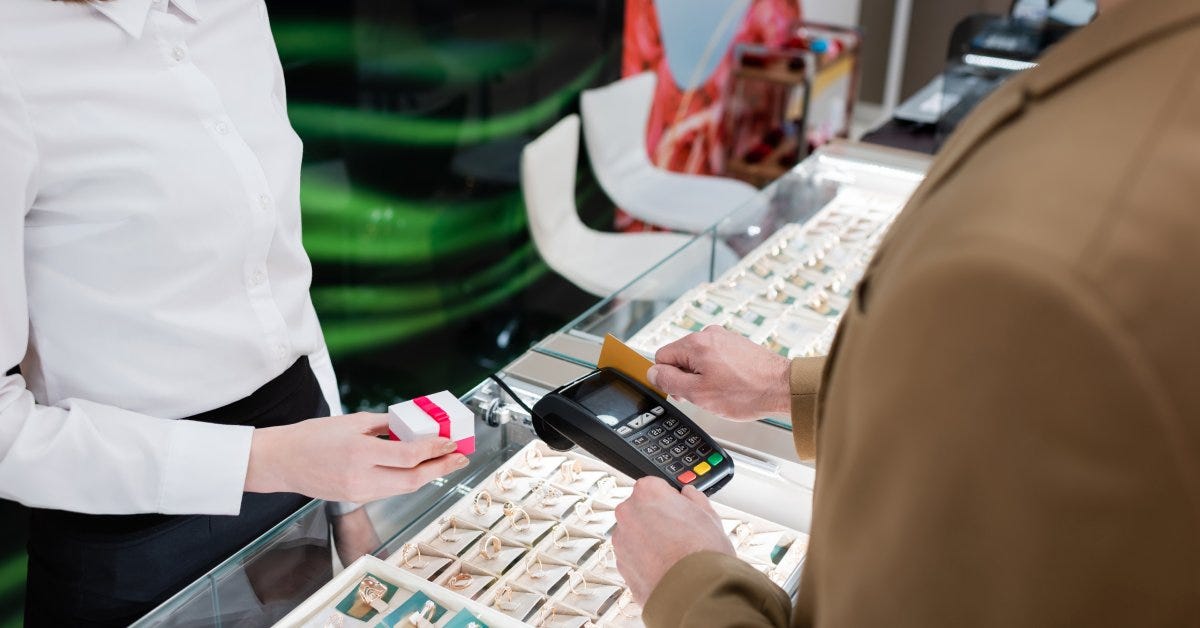 A seller at a jewelry store holds a small gift box wrapped in red ribbon as a customer pays for the item.