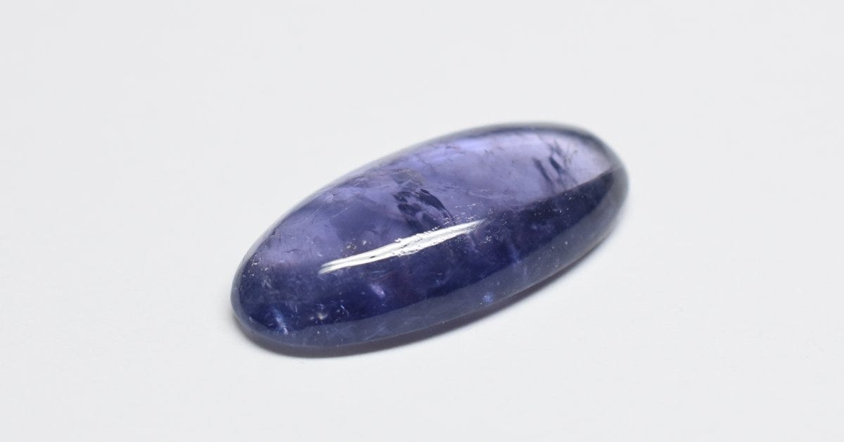A cabochon-cut iolite gemstone rests on a white surface. Its purple hue shines, showcasing its polished finish.