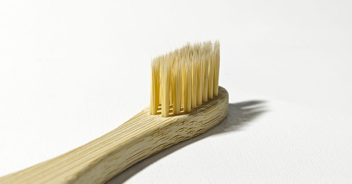 A close-up of a toothbrush with a wooden handle against a white background, illuminated by light coming from the left.