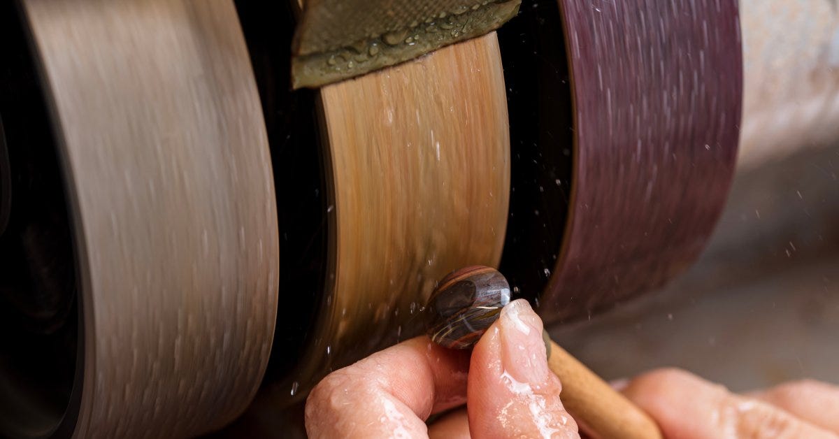 A lapidary holds a small tiger's eye gemstone against a grinding wheel and uses wet sanding to polish its surface.