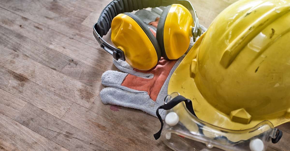 Safety gear, including a yellow hard hat, noise-canceling earmuffs, eye protection, and gloves, laid out on a wooden table.