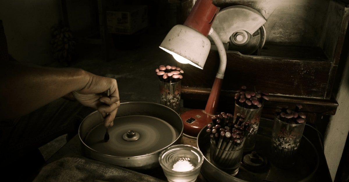 Hand holding a dop stick over a lapidary machine inside a home workshop with multiple dop sticks in glasses next to a desk lamp.