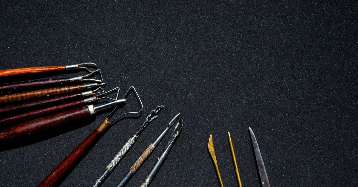 Multiple wax carving tools of different sizes and shapes with metal tips lined up next to each other placed over a gray surface.