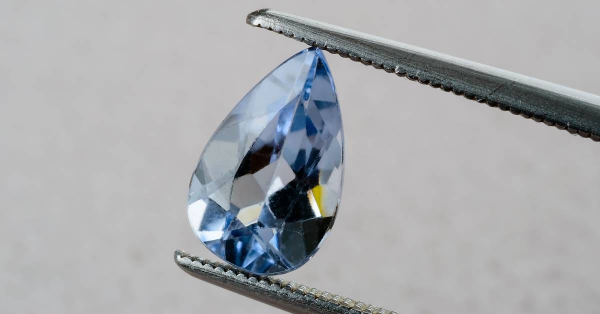 Close-up to a light blue gem with multiple facets held in between a pair of lapidary tweezers with a gray wall background.