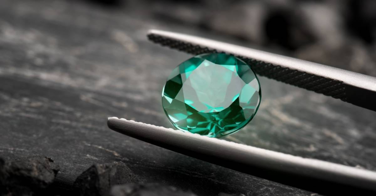 Close-up to a green gem with a rose cut held in between a pair of lapidary tweezers with a dark rock in the background.