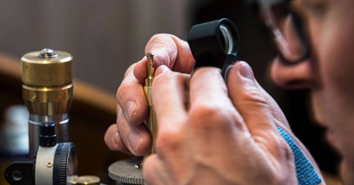 A man wearing glasses, holding a jewelry magnifying glass close to a small stone. He holds the stone with a metal dupe.