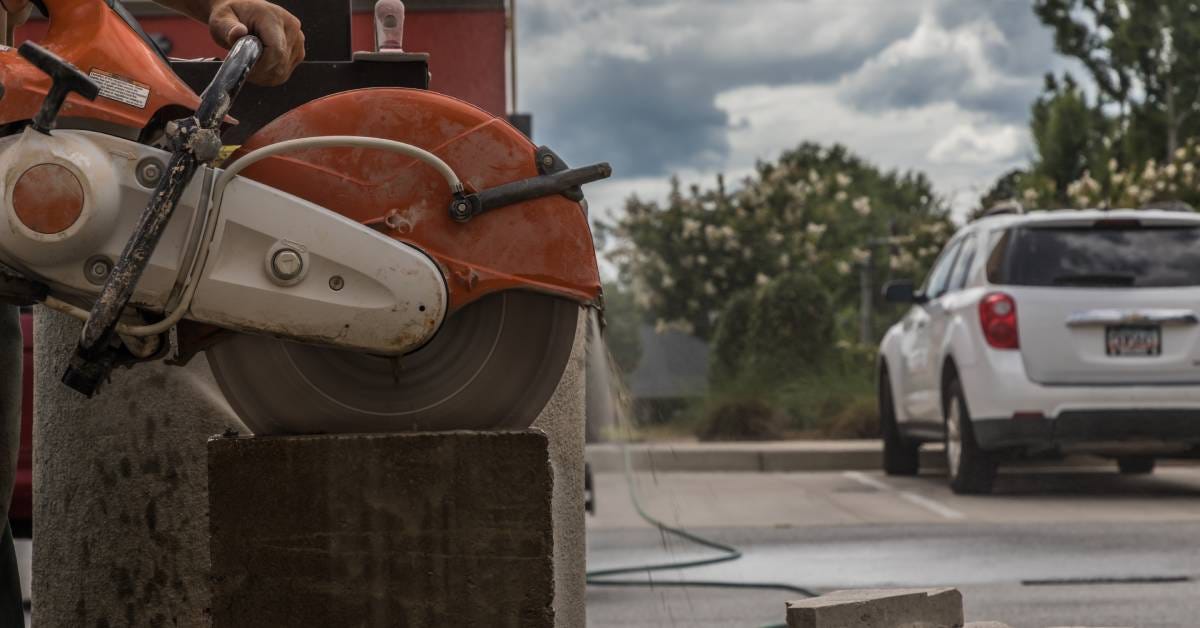A man holding a large orange stone grinder with a large blade. He cuts a large cube of stone outdoors.