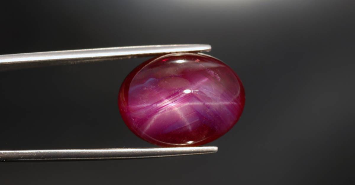 A pair of tweezers holding a small shiny red ruby with a smooth cabochon cut. A white light brightens the stone.