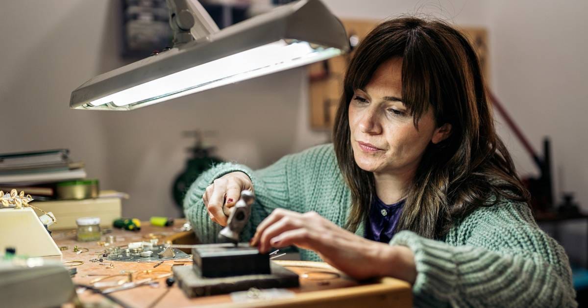 Woman wearing green cardigan sweater holding a hammer while working on jewelry pieces under a bright overhead lamp.